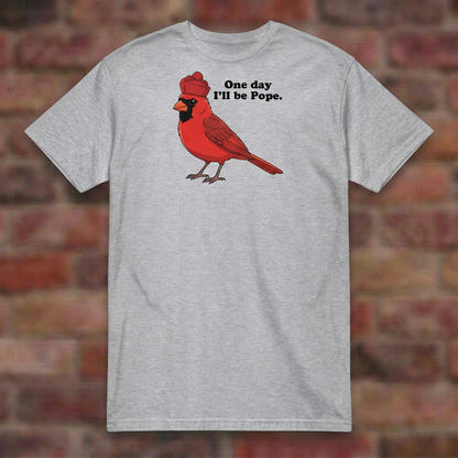 One day I'll be Pope T-shirt in Sport Grey, flat lay on blurred brick